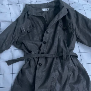 Grön jacka  - Grön jacka från Micha med knappar framtill, bälte i midjan och coola detaljer med nitar på bröstfickorna. Den är lång ärmar och passar perfekt till våren eller sommaren 