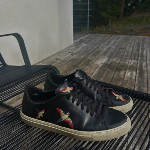 Svarta sneakers med broderade fåglar - Svarta sneakers i skinn från Axel Arigato med broderade fåglar i rött, blått och beige på sidorna och tån. Klassisk låg modell med svarta snören och vit sula. Perfekta för dig som vill ha en unik detalj på fötterna.