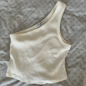 Vit ribbad oneshoulder topp NA-KD - Säljer en stilren vit ribbad topp från NA-KD i oneshoulder-modell. Toppen är ärmlös och har en tight passform som framhäver axeln. Perfekt för att skapa en trendig look till sommaren. Tveka inte att höra av dig!💗