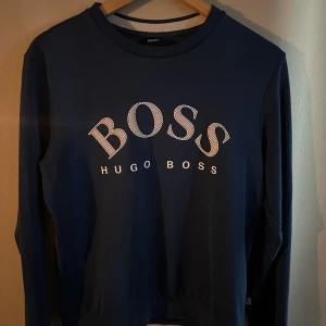 Blå långärmad tröja från Hugo Boss - Snygg blå långärmad tröja från Hugo Boss med stort vitt BOSS-tryck på bröstet. Klassisk rund halsringning och mjukt material som känns skönt mot huden. Perfekt för dig som gillar stilrena och trendiga plagg med tydlig logga.