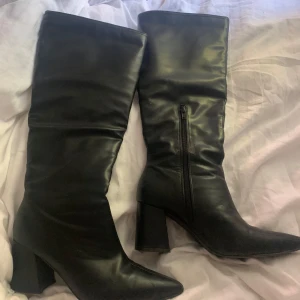 Svarta höga boots med klack - Snygga svarta boots i skinnimitation med spetsig tå och 7 cm blockklack. Stövlarna har dragkedja på insidan för enkel påtagning och en stilren, minimalistisk look. Perfekta för att lyfta vilken outfit som helst. Lite sletna vid dragkedjan men funkar felfritt. Från Nelly. Köpta för 800. St 38
