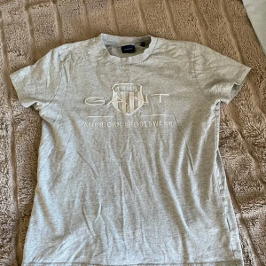 Grå GANT t-shirt med broderad logga - Snygg grå t-shirt från GANT med broderad logga och texten 'American Sportswear' framtill. Klassisk rund hals och korta ärmar. Tillverkad i mjuk bomull för en skön känsla. Perfekt för en avslappnad och sportig stil.