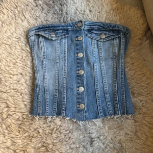 Jeanskorsett från Zara med knappar - Säljer en snygg jeanskorsett från Zara i ljusblå denim. Toppen har knappar framtill, dekorativa ficklock och rå, fransig nederkant. Baksidan har smock för extra stretch och passform. Perfekt att styla med höga byxor eller kjol.