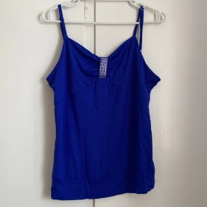 Blå linnetopp med strassdetalj - Snygg blå linnetopp med smala justerbara axelband och en glittrig strassdetalj framtill. Toppen har en lätt rynkad effekt vid bysten och är tillverkad i mjukt bomullsmaterial. 