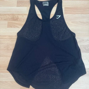 Svart linnetopp från Gymshark - Säljer en svart, luftig linnetopp från Gymshark med brottarrygg och diskret logga framtill. Toppen är tillverkad i 100% polyester och har en lätt transparent känsla, perfekt för träning. Snyggt rundad nederkant och sportig passform.