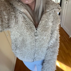 Beige teddyjacka med huva - Säljer en fluffig beige teddyjacka med huva och dragkedja framtill. Jackan har en oversized look, ribbade muddar och ett stort ringformat dragkedjehandtag. Perfekt för dig som gillar mysiga och trendiga jackor med cool vibe.