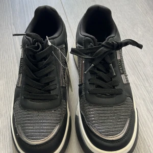 Svarta sneakers med metallicdetaljer - Snygga svarta sneakers från Melton med coola metallicdetaljer på sidorna och hälen. Ovandelen har ett randigt mönster i svart och silver, och skorna har tjock vit sula för extra komfort. Klassisk snörning och komfortkudde för skön känsla hela dagen. Ej använd helt ny. 