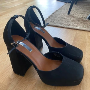 Svarta platåklackar - Säljer ett par svarta platåklackar från Primark med fyrkantig tå och justerbart ankelband. Skorna har 9,5 cm hög blockklack och plattform. Materialet är matt syntet och de passar perfekt till jeans eller klänning. Använda en gång.