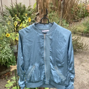 Blå bomberjacka från Wera Stockholm - Snygg blå bomberjacka från Wera Stockholm i storlek 34. Jackan har dragkedja framtill, ribbade muddar och cool rynkdetalj längs ryggen och ärmarna. Materialet känns lätt och har en silkeslen finish. Perfekt för dig som vill ha en stilren men unik jacka.