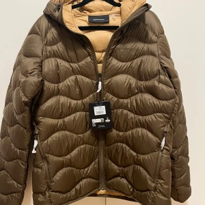 Brun dunjacka från Peak Performance - Snygg brun dunjacka från Peak Performance med vågformat quiltat mönster. Jackan har huva, dragkedja framtill och två sidofickor med dragkedja. Insidan är beige och materialet är lätt och smidigt, perfekt för kyliga dagar. Ordinariepris är 3000. 