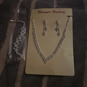 Glittrande smyckeset Forever Victory - Snyggt smyckeset med halsband, armband och långa örhängen i silverfärg. Alla delar är täckta av glittrande stenar i ett mönster som ger extra bling. Perfekt för dig som vill sticka ut med en lyxig och glittrig look.