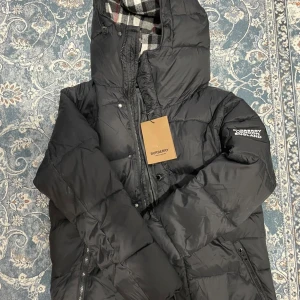 Svart pufferjacka från Burberry - Svart pufferjacka från Burberry med stor huva och quiltad design. Jackan har dragkedja framtill, Burberry-logga på ärmen och rutigt foder i huvan. Perfekt för kalla dagar och har en riktigt clean look.