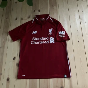 Liverpool FC röd matchtröja New Balance - Liverpool FCs officiella matchtröja från New Balance i rött med vita detaljer och tryck. Kortärmad med krage och knappar, tillverkad i lätt och ventilerande polyester. Tryck med Standard Chartered och Western Union på bröst och ärm, samt LFC-logga broderad.