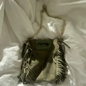 Olivgrön mocka axelväska från Maje - Säljer en olivgrön axelväska från Maje i mocka med fransar på sidorna och kedjerem. Väskan har ett grönt handtag upptill och medföljer även en extra axelrem. Perfekt för dig som vill ha en unik och trendig accessoar. 💞💞Längre rem och dustbag ingår🥰