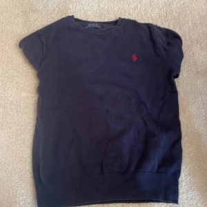 Mörkblå tröja från Polo Ralph Lauren - Mörkblå tröja från Polo Ralph Lauren med röd broderad logga på bröstet och bruna manchesterlappar på armbågarna. Klassisk rund halsringning och långa ärmar med ribbade muddar. Perfekt för dig som gillar stilrena detaljer.