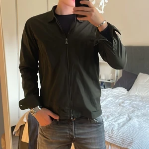 Mörkgrön overshirt från Oscar Jacobson - Stilren mörkgrön overshirt från Oscar Jacobson med dragkedja framtill, klassisk krage och bröstficka. Jackan är i ett lätt bomullsmaterial och har långa ärmar samt justerbara knappar i nederkant. Perfekt för lager-på-lager och snygg till jeans.