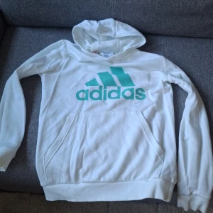 Vit hoodie från Adidas med turkost tryck - Vit hoodie från Adidas med stort turkost logotryck på bröstet. Klassisk känguruficka framtill, ribbade muddar och huva. Mjuk bomull. OBS finns liten färgfläck på insidan av muddarna nedtill, ingenting som syns utanpå direkt men annars i fint skick, pruta om priset inte funkar annars klicka hem för 100 spänn