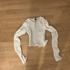 Vit smockad blus med volangkant - Supersöt vit blus från gina med smockad kropp och långa, lätt transparenta ärmar. Toppen har volangkant nertill och en rund halsringning. Perfekt för dig som gillar romantisk och trendig stil.