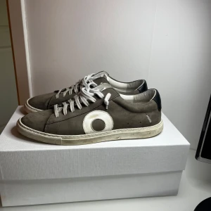 Oliver Cabell Low 1 Smoke sneakers 41 - Snygga sneakers från Oliver Cabell i modellen Low 1, färg smoke. Skorna har en grå mocka-överdel med vit rund detalj på sidan, vita snören och svart hälparti. Klassisk låg profil och platt vit sula. Perfekta för dig som gillar stilrena och trendiga sneakers.