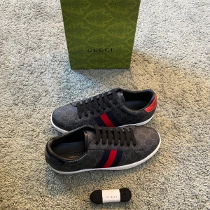 Gucci ace skor - Snygga Gucci sneakers i mörkgrått med klassiskt GG-mönster, röda och blå ränder på sidan samt olika färg på hälarna – en röd och en blå. Skorna har svarta skosnören, vit sula och detaljer i skinn. Kommer med originalkartong och extra skosnören.