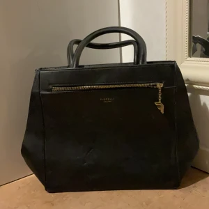 Svart handväska från Fiorelli - Stilren svart handväska från Fiorelli med guldfärgade detaljer och dragkedja framtill. Väskan har två rejäla handtag och en rymlig form, perfekt för att bära med sig allt man behöver. Materialet är slätt och ger ett klassiskt intryck.