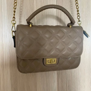 Quiltad beige axelväska - Snygg quiltad axelväska i beige skinnimitation med guldfärgade kedjedetaljer och vridlås. Väskan har både handtag och lång axelrem, samt flera fack inuti med dragkedja. Perfekt för dig som vill ha en stilren och trendig väska med lyxig känsla.