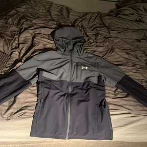 Under Armour grå vindjacka med huva - Snygg tvåfärgad vindjacka från Under Armour i mörkgrått och ljusgrått. Jackan har huva, dragkedja hela vägen, två sidofickor och en bröstficka med dragkedja. 