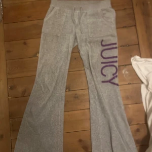 Grå Juicy Couture mjukisbyxor bootcut - Grå mjukisbyxor från Juicy Couture med lila glittrig JUICY-text längs benet. Byxorna har bootcut-modell, låg midja, snörning i midjan och fickor både fram och bak. Fickorna där bak är med den gamla designen! Perfekta för en chill och trendig look.