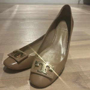 Beige pumps från Tory Burch  - Snygga beige pumps från Tory Burch i glansigt skinn med rund tå och låg klack. Framtill sitter en stor guldig logodetalj som ger skorna en lyxig touch. Perfekta för dig som vill ha en klassisk men trendig look. ÄKTA!! 