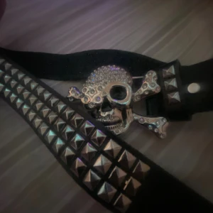Svart nitarbälte med dödskalle-spänne - Säljer ett svart bälte med coola silverfärgade nitar och ett stort spänne i form av en glittrig dödskalle med korslagda ben. Perfekt accessoar för dig som vill sticka ut och ge din outfit en edgy vibe. Bältet är brett och har tydliga punkinfluenser. Tar Bara seriösa köpare.