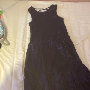 Glittrig marinblå långklänning Lindex - Supersnygg marinblå långklänning från Lindex med glittrigt tyg och coola korsade band i ryggen. Klänningen är ärmlös och har en lätt utställd kjol som ger en riktigt elegant vibe. Perfekt för dig som vill sticka ut med lite extra glitter!