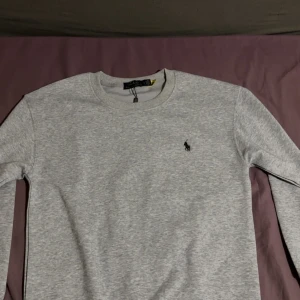 Grå tröja från Polo Ralph Lauren - Snygg grå tröja från Polo Ralph Lauren med klassisk rund halsringning och broderad logga på bröstet. Tröjan är långärmad och har en stilren, enkel design som passar perfekt till jeans eller chinos.