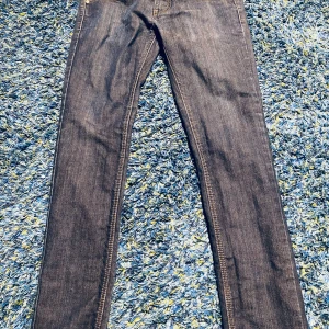 Mörkblå jeans från Nudie Jeans - Säljer ett par mörkblå jeans från Nudie Jeans med klassiska orange sömmar och broderade bakfickor. Jeansen har rak passform och är tillverkade i slitstark denim av bomull. Perfekta för dig som gillar stilrena och tidlösa jeans.