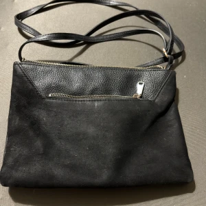 Svart axelväska i skinn och mocka - Snygg svart axelväska med lång rem, kombinerat i slätt skinn och mocka. Väskan har en dragkedja upptill och en extra ficka med dragkedja framtill. Perfekt för dig som gillar stilrena accessoarer med lite edge.