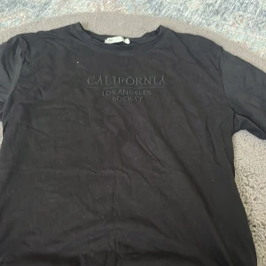 Svart t-shirt från Fullamoda med tryck - Svart t-shirt från Fullamoda med texten 'California Los Angeles Society' tryckt framtill. Klassisk rund halsringning och korta ärmar. Skön och enkel modell i bomull, perfekt till jeans eller shorts. Ser jätte skrynklig ut i bilden men är inte det säljer den på grund av att den var för stor