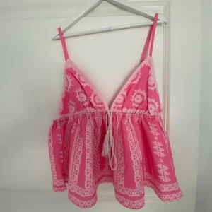 Rosa mönstrad linnetopp med knyt - Supersöt rosa linnetopp med vitt etnoinspirerat mönster och smala axelband. Toppen har v-ringning, justerbara axelband och knyt i midjan med tofsar. Luftig och flowy passform, perfekt för varma dagar. Den är från en marknad i Spanien.