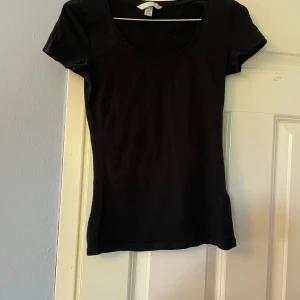 Svart kortärmad topp från H&M - En enkel svart topp från H&M i storlek XS. Modellen har rund halsringning och korta ärmar, perfekt för en clean och stilren look. Tillverkad i mjuk bomull som känns skön mot huden och passar till många olika outfits.