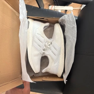 Vita Adidas Ultraboost sneakers - Säljer ett par fräscha vita Adidas Ultraboost sneakers med stickad ovandel och klassiska tre ränder på sidorna. Skorna har snörning, platt sula och Boost-dämpning för extra komfort. Perfekta för dig som gillar stilrena och sportiga skor.               MOTIVERING: helt nya ultraboost som endast är använda en enda gång påväg till Ica och tillbaka. De är i nytt skick och luktar nya sneakers. Skorna var alldeles för stora så säljer de här på ca 50% rea