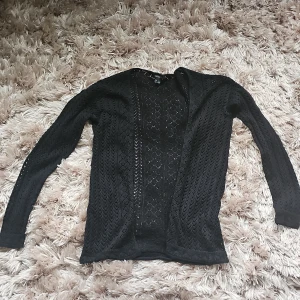 Svart stickad kofta från Esmara - Svart, tunn och stickad kofta från Esmara i storlek S. Koftan har ett snyggt hålmönster över hela plagget och är långärmad. Perfekt att slänga över en topp för en chill look. Materialet är mjukt och luftigt.