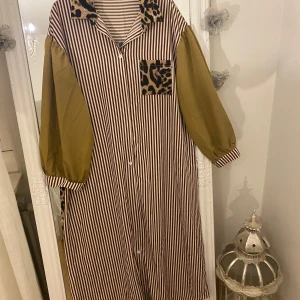 Randig långklänning med leoparddetaljer - Unik långklänning med bruna och vita vertikala ränder, kontrasterande olivgröna ballongärmar och coola leopardmönstrade detaljer på krage, bröstficka och rygg. Knäppning framtill och bred passform. Perfekt för dig som gillar statement-plagg och mixade mönster.