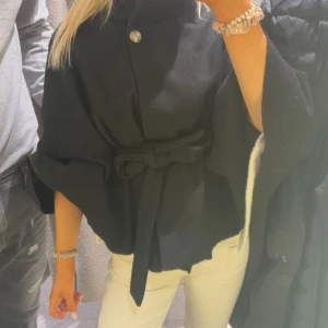 Svart kappa  - Säljer en svart kappa med bälte i midjan och en snygg knapp upptill. Jackan har vida ärmar och är i ett mjukt tyg som känns lyxigt. Perfekt för dig som vill ha en stilren och trendig look. Från missäy