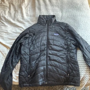 Svart quiltad dunjacka från Patagonia - Svart dunjacka från Patagonia med quiltad design och hög krage. Jackan har dragkedja framtill och två sidofickor med dragkedja. Perfekt för kyliga dagar och enkel att matcha med allt. Klassisk logga på bröstet.