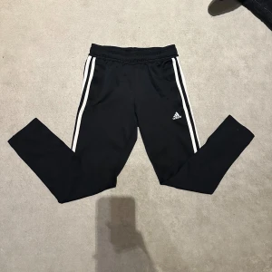 Svarta Adidas träningsbyxor med vita ränder - Svarta träningsbyxor från Adidas med klassiska vita ränder längs sidorna och Adidas-logga på låret. Byxorna har elastisk midja och smal passform, perfekta för sport eller chill. Tillverkade i mjukt och stretchigt syntetmaterial.