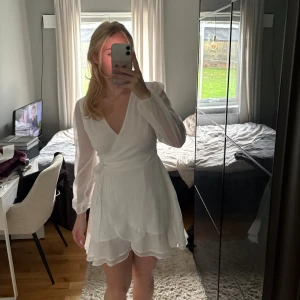 Vit omlottklänning med volang - Superfin vit kort omlottklänning med långa, lätt transparenta ärmar och volangkant nertill. Klänningen har v-ringning och knyt i midjan för en snygg passform. Perfekt för sommarens alla tillfällen!