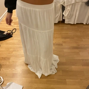 Vit långkjol med volanger - Säljer en vit långkjol med bred resår i midjan och snygga volangdetaljer. Kjolen har ett flowigt fall och är tillverkad i ett lätt, vävt material som känns luftigt och skönt. Perfekt för dig som gillar en bohemisk och chill stil. Vet tyvärr inte vart jag köpr den kom med bud eller skriv om ni har frågor jag tömmer min garderob så gärna snabbt köp!