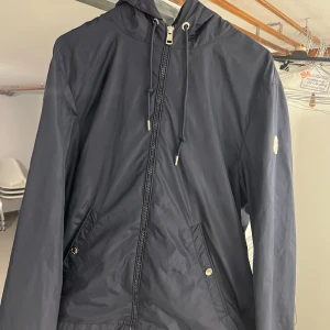 Svart vindjacka från Moncler - Snygg svart vindjacka från Moncler med huva och coola snörningsdetaljer. Jackan har dragkedja framtill, fickor med tryckknappar och Moncler-logga på ärmen. Materialet är lätt och syntetiskt, perfekt för blåsiga dagar. Röd och vit detalj på huvan ger extra stil.