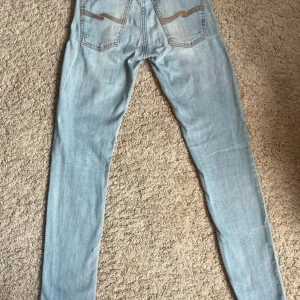 Ljusblå skinny jeans från Nudie Jeans - Säljer ett par ljusblå skinny jeans från Nudie Jeans med klassisk orange söm på bakfickorna och diskret logga. Jeansen har hög midja och är tillverkade i mjuk bomullsdenim. Perfekta för dig som gillar en tight passform och stilren look.