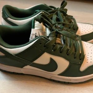 Nike Dunk Low gröna och vita sneakers - Nike Dunk Low sneakers i grönt och vitt skinn med klassisk swoosh på sidan. Lågt skaft, gröna snören och perforerad tå för extra ventilation. Snygg kontrast mellan de gröna och vita panelerna, perfekt för dig som gillar streetstyle och sportig look.