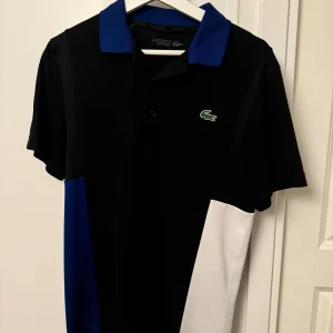 Lacoste Sport svart/blå/vit piké S - Snygg pikétröja från Lacoste Sport i svart, blått och vitt. perfekt för en sportig och stilren look. Logga på ärmen och ryggen.