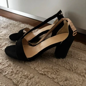 Svarta klackar strl 38 - Stilrena svarta klackar. Skorna har en enkel rem över tårna och justerbart spänne vid ankeln i guldton. Tillverkade i mockaliknande material och passar perfekt när du vill lyfta din outfit. Aldrig använda då dem va för små för mig